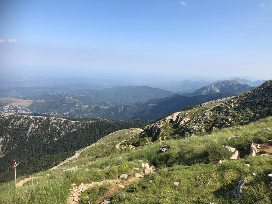 Looking back down a grassy slope with narrow rocky path visible, and a signpost in the bottom left of the picture. Looking further afield, there are slopes covered by woodland, and mountain ranges on the horizon.