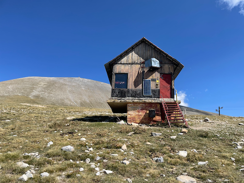 Mountain hut on raised platform, with rounded hillside behind.