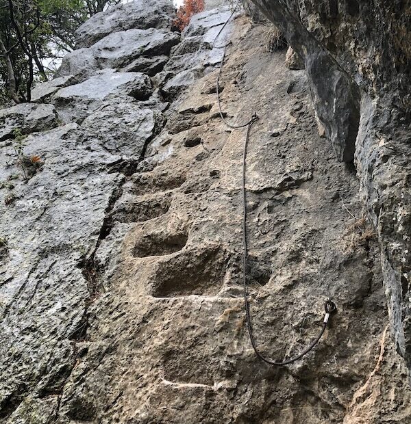 View from below of ancient rock-cut steps on cliff face with fixed metal rope beside them.