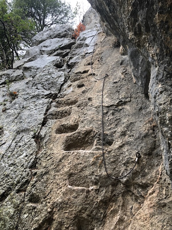 View from below of ancient rock-cut steps on cliff face with fixed metal rope beside them.